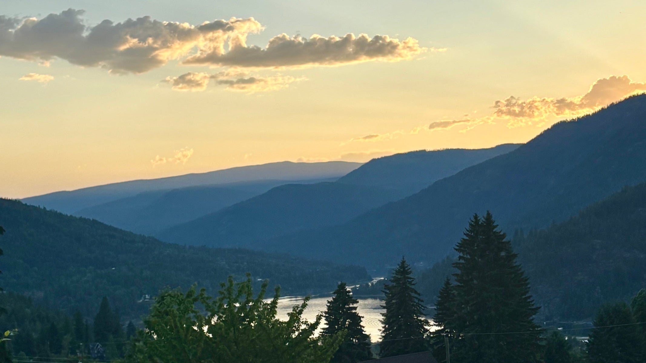 Scenic view of mountains and a lake at sunset with trees and buildings in the foreground.