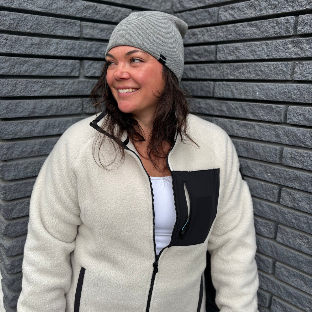 Woman wearing a white jacket with black accents and a gray beanie standing against a gray brick wall.