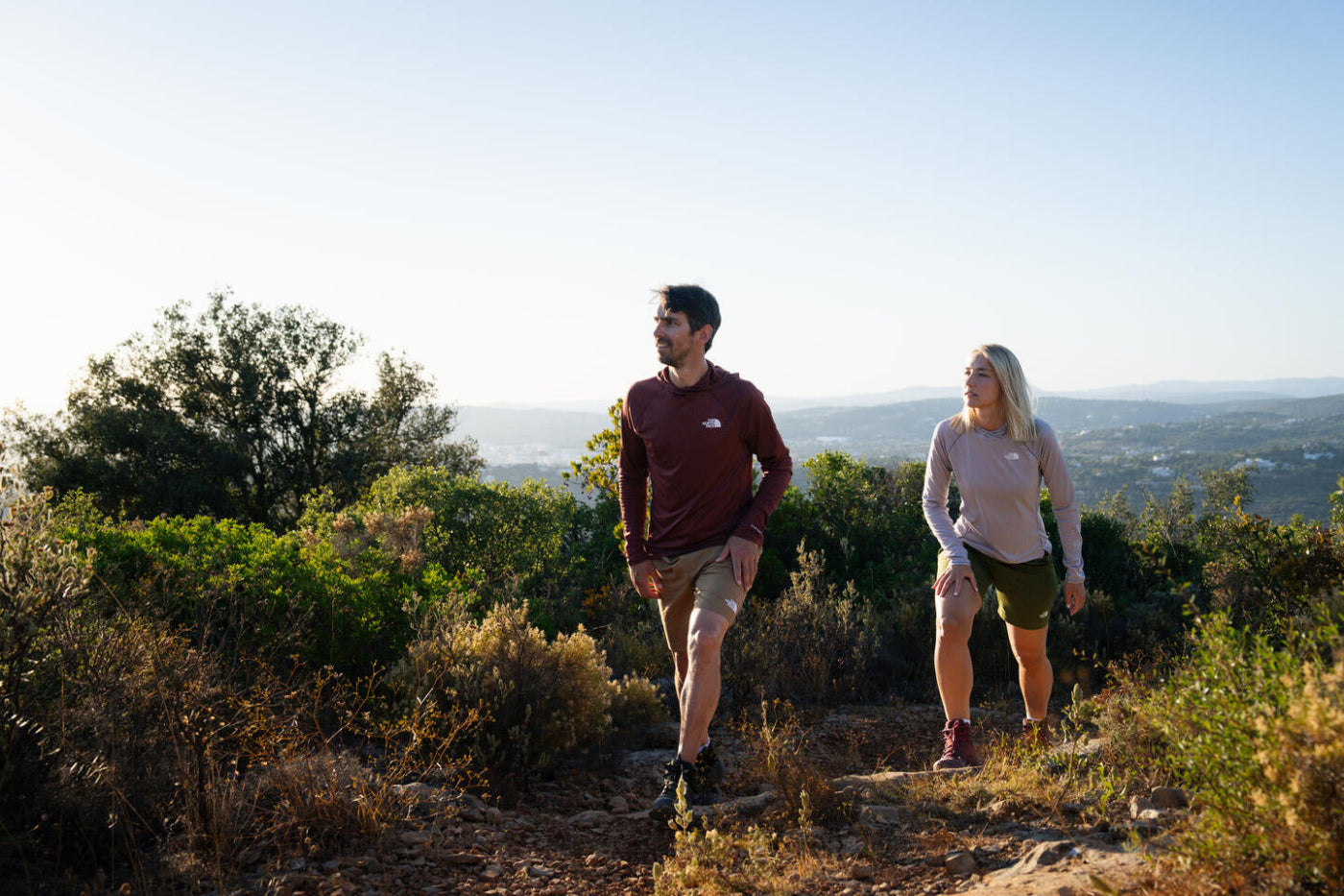 A man and a woman hiking on a sunlit trail in a mountainous outdoor setting, wearing matching long-sleeve athletic shirts in dark red and light pink.
